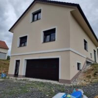 Nově postavený dům se žlutou fasádou, tmavě hnědými okny a dveřmi, doplněný o vjezd do garáže